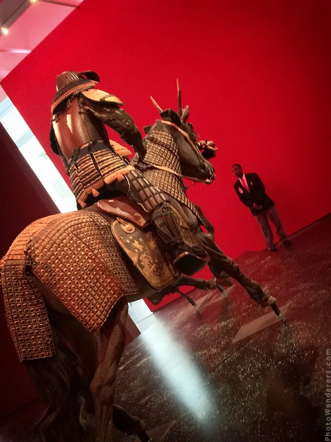 Horse armor, rear view, against the red wall. The rider leans forward. The whole display fills the room.