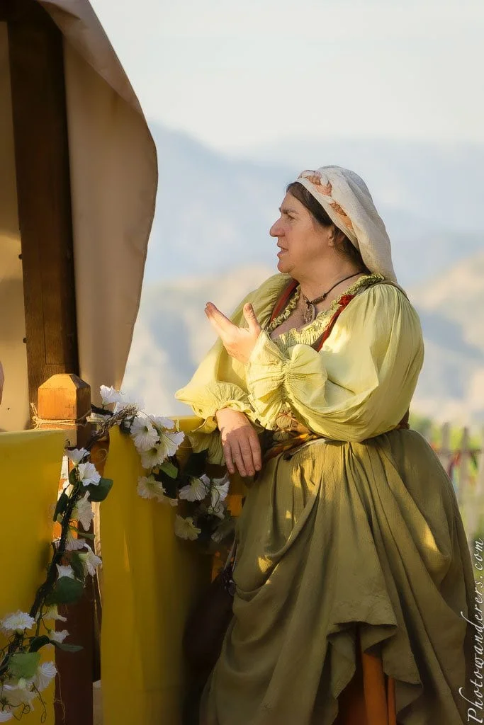 A village woman leans over the fence, mid-conversation, flowers tumbling beside her. 