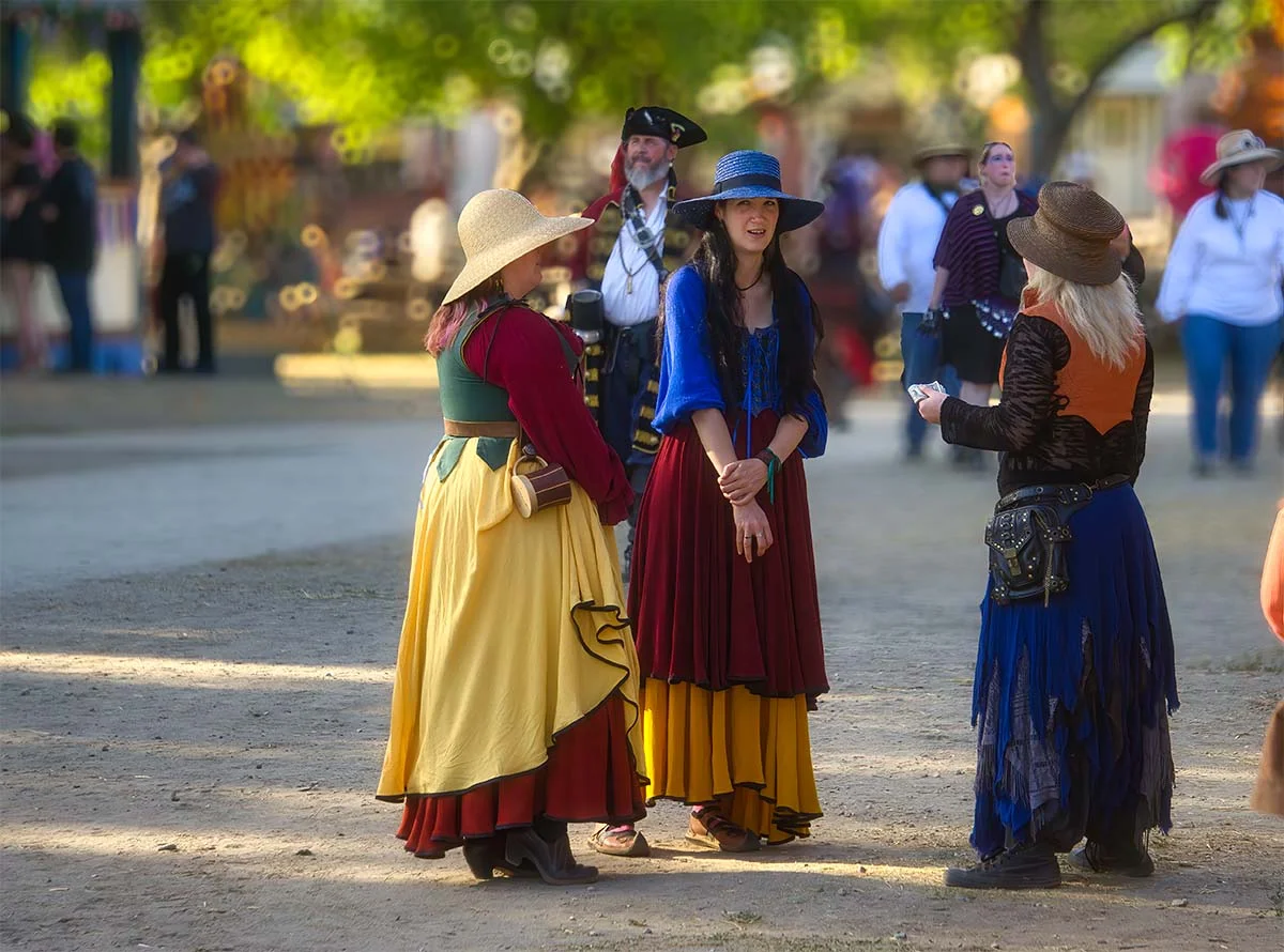 Yellow, crimson, cobalt blue. Three women mid-conversation on a dusty street. 
