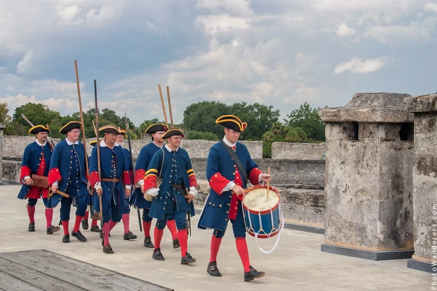 Colonial reenactors in period uniforms marching on fort ramparts with drum